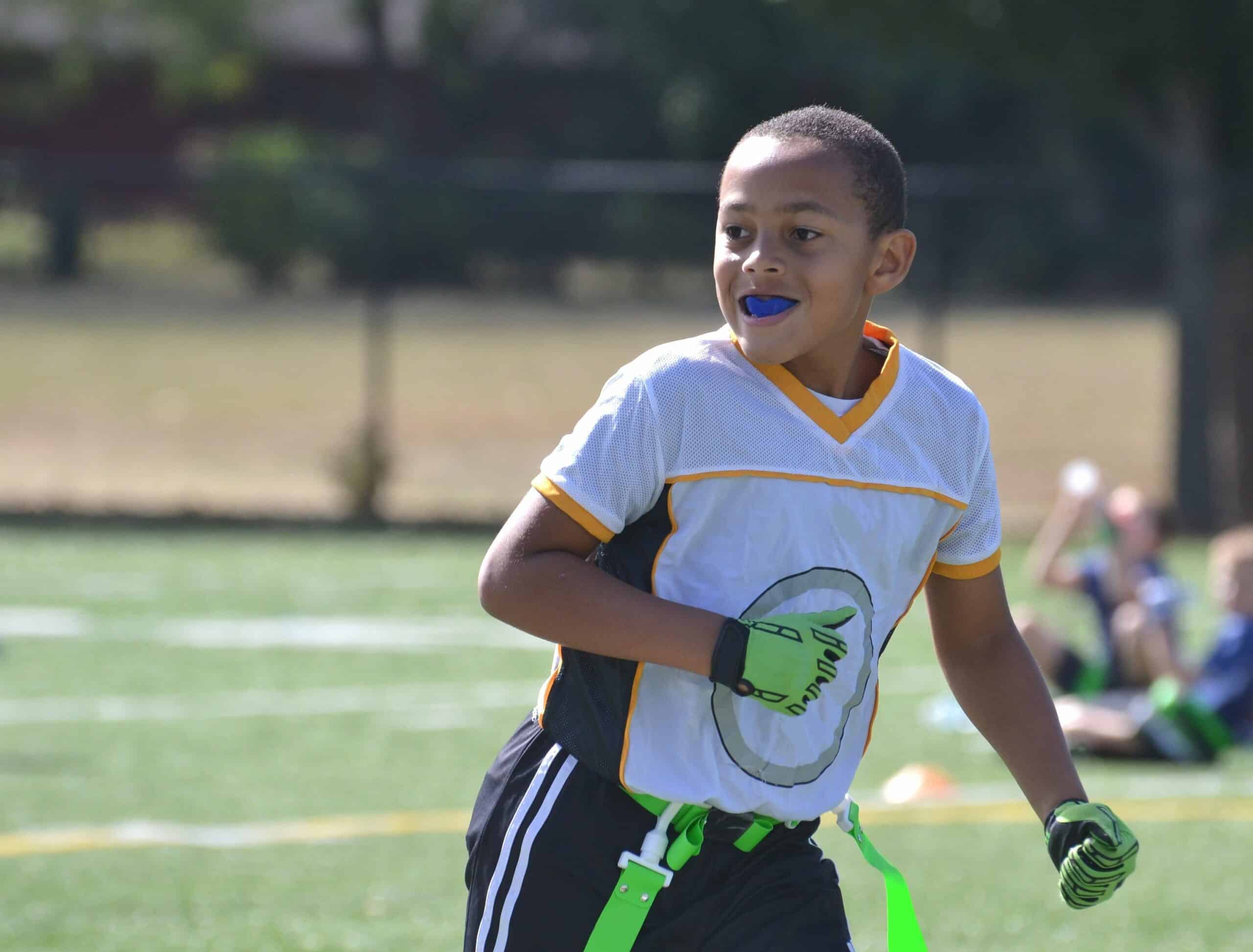 A child wearing a sports jersey and mouthguard runs on a grassy field, surrounded by trees in the background, with others seated nearby.