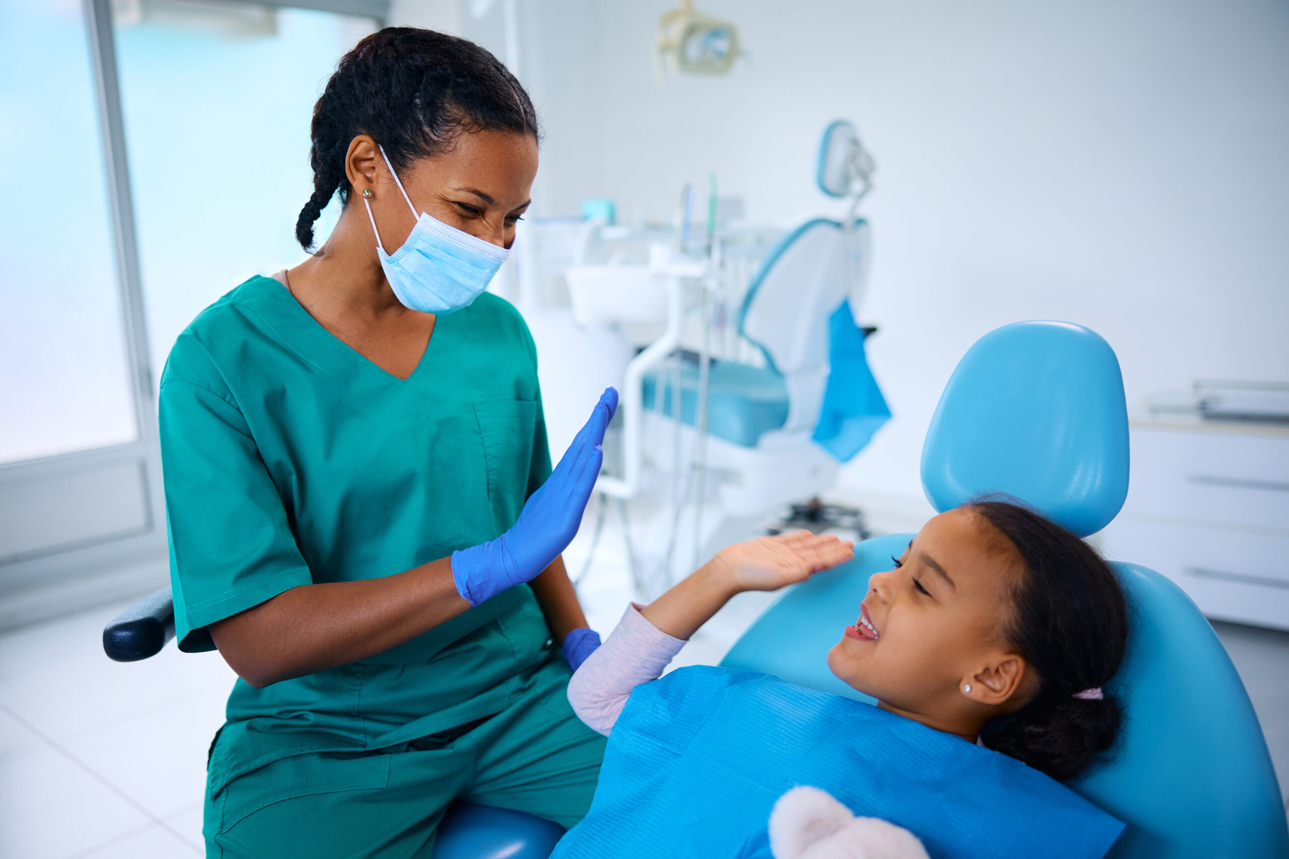 A person in scrubs high-fives a child in a dental chair, both smiling in a bright, modern dental office setting.