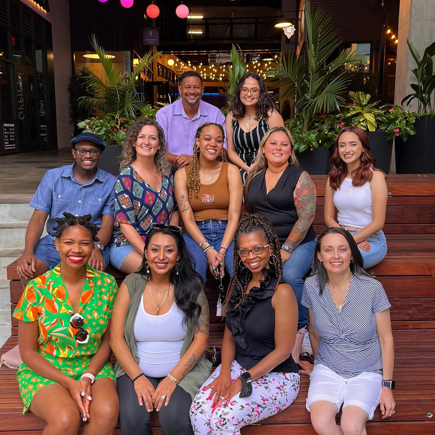 A group of eleven people smiling, sitting on wooden steps surrounded by greenery and colorful lights, outside an urban venue.
